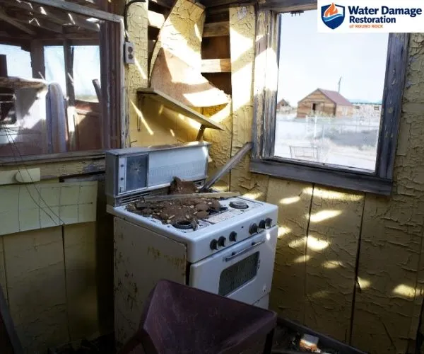 view of a fire-damaged kitchen showing a debris-covered stove, peeling yellow wall paint, and exposed wall framing.